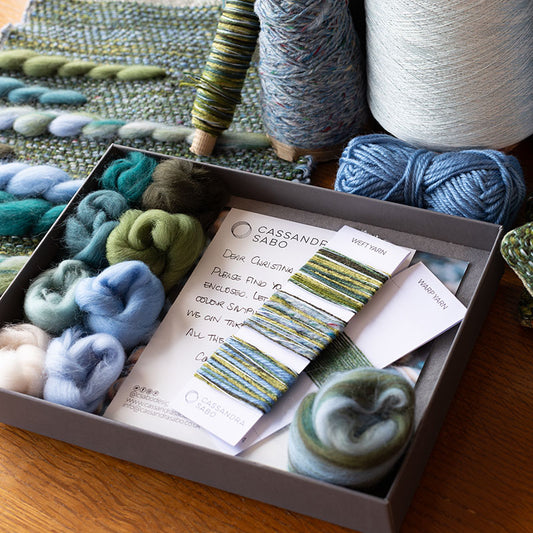 Desk with handwoven samples and a grey box filled with yarn and fibre samples and yarn windings for a comission