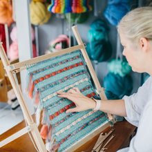 Load image into Gallery viewer, Cassandra Smith sitting at her desk and weaving a cushion cover on The Oxford Frame Loom with a backdrop of colour wool roving in macrame planter holders