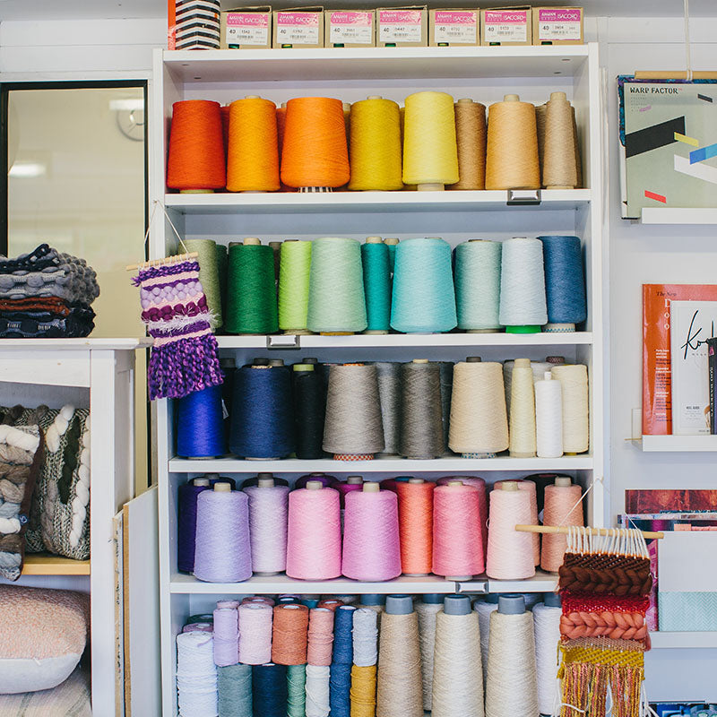 A shelf filled with colourful 2/16s mercerised cotton yarn cones surrounded by weaving books and handwoven products